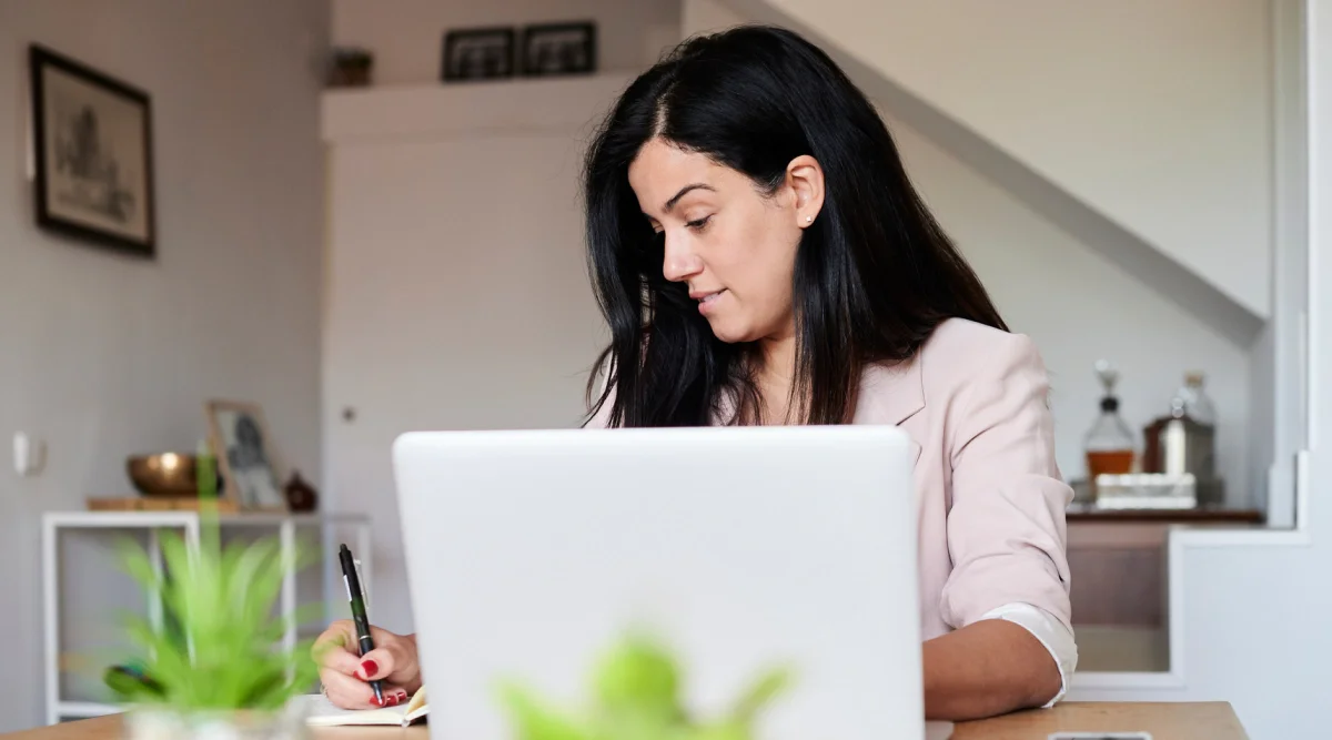 A woman writes in a notebook while focused on her laptop in a modern office setting