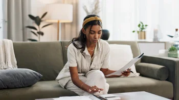 A woman reviews papers while sitting on a couch, with a smartphone nearby in a bright living room