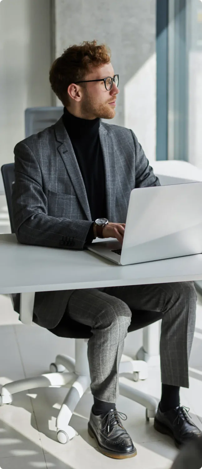 White man with red hair, glasses and a 5 o'clock shadow  wearing a grey suit with a black turtle neck sitting in the office with a white laptop filling out his business dissolution with LegalZoom.