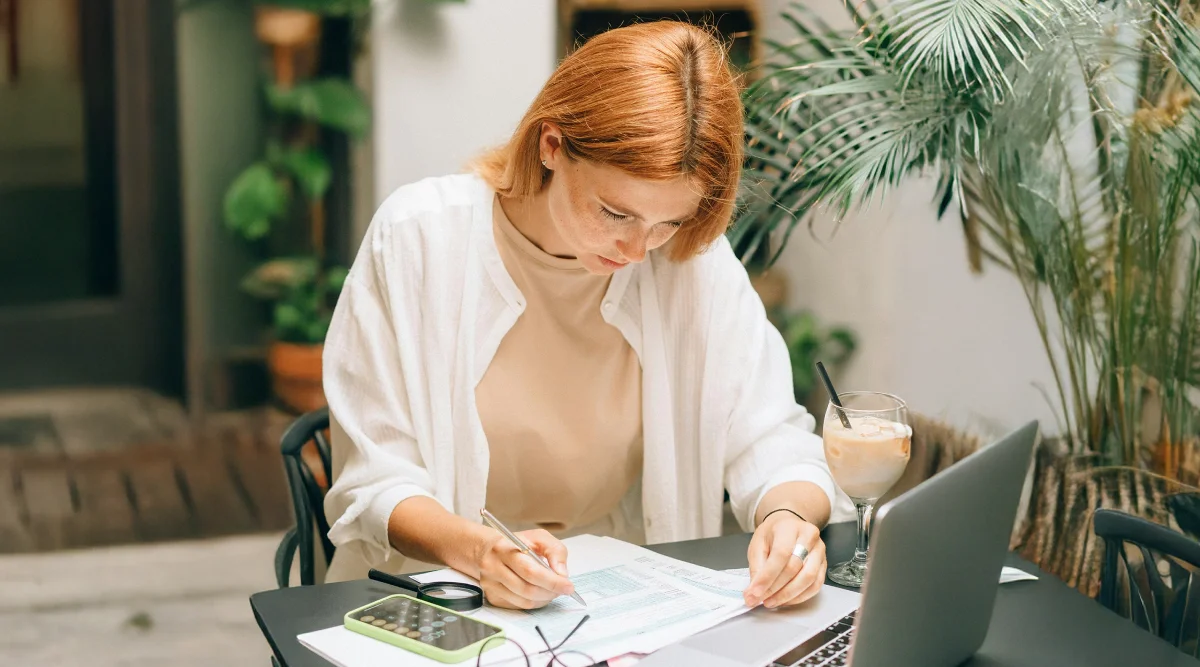 A woman writes notes at a sunny outdoor cafe with a laptop and a drink beside her