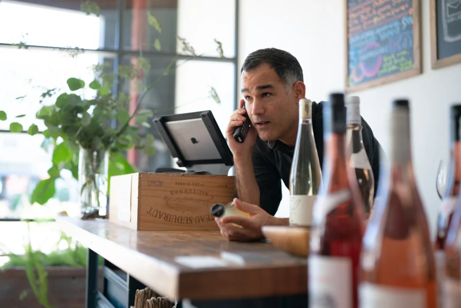 White man holding a telephone to his ear looking off into the distance at the cash register. Wine bottles in the foreground.