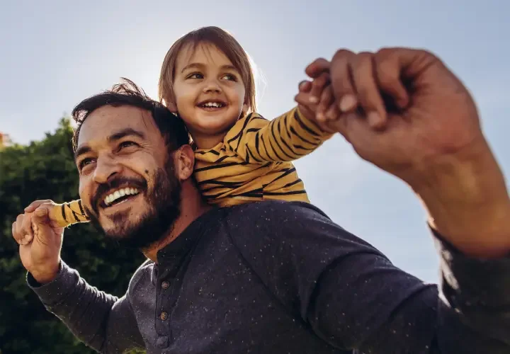 A smiling father carries his daughter on his shoulders. The daughter is spreading her arms out in flight.