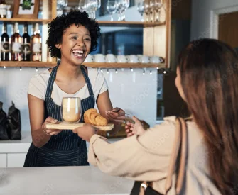 Black woman with curly hair wearing a white t-shirt and a blue and white pinstriped apron working in a café and  smiling because she got her business license from LegalZoom as she hands a woman with brown hair wearing a tan shirt her order.