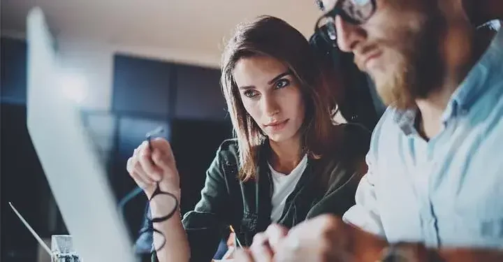 Close-up of a man and a woman sitting at a desk and looking over the same laptop.