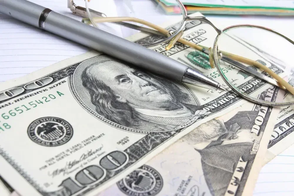 A pen and a pair of wire-rimmed glasses sit atop a stack of money.