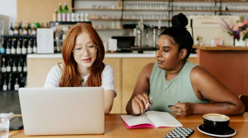 Two women collaborating in a café with a laptop and notes