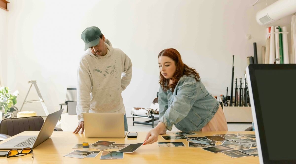 A man and a woman stand in a sunny photography studio and sort through prints of photos.