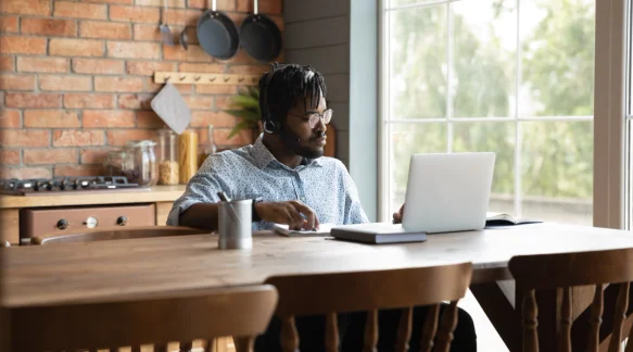 A black man wearing glasses sits at his desk and uses a laptop.