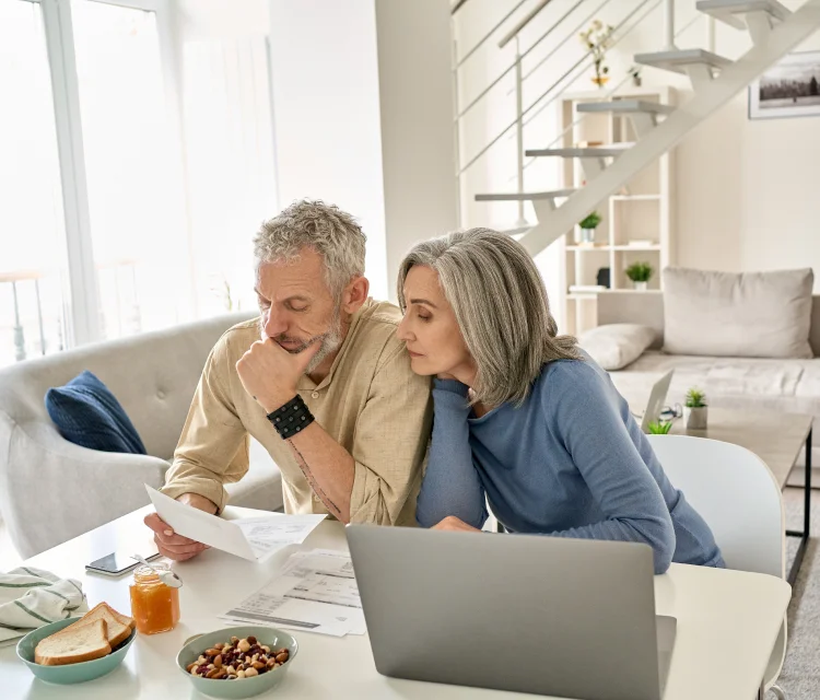 a man with white hair and a beard holds a bill thoughtfully, while a woman with white hair looks over his shoulder.