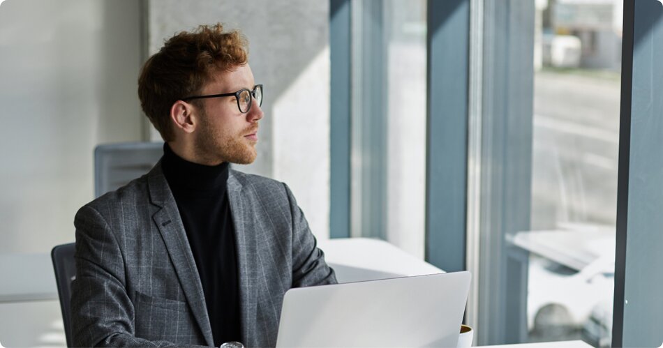 White man with red hair, glasses and a 5 o'clock shadow  wearing a grey suit with a black turtle neck sitting in the office with a white laptop filling out his business dissolution with LegalZoom.