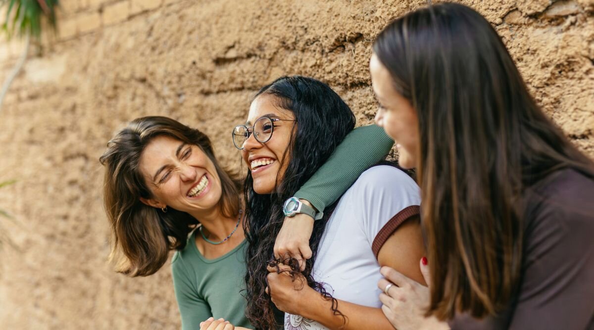 Two women embrace their teenage daughter as all three laugh.