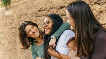 Two women embrace their teenage daughter as all three laugh.