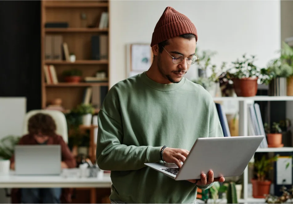 A man in a brown beanie and glasses holds his laptop and types one handed.