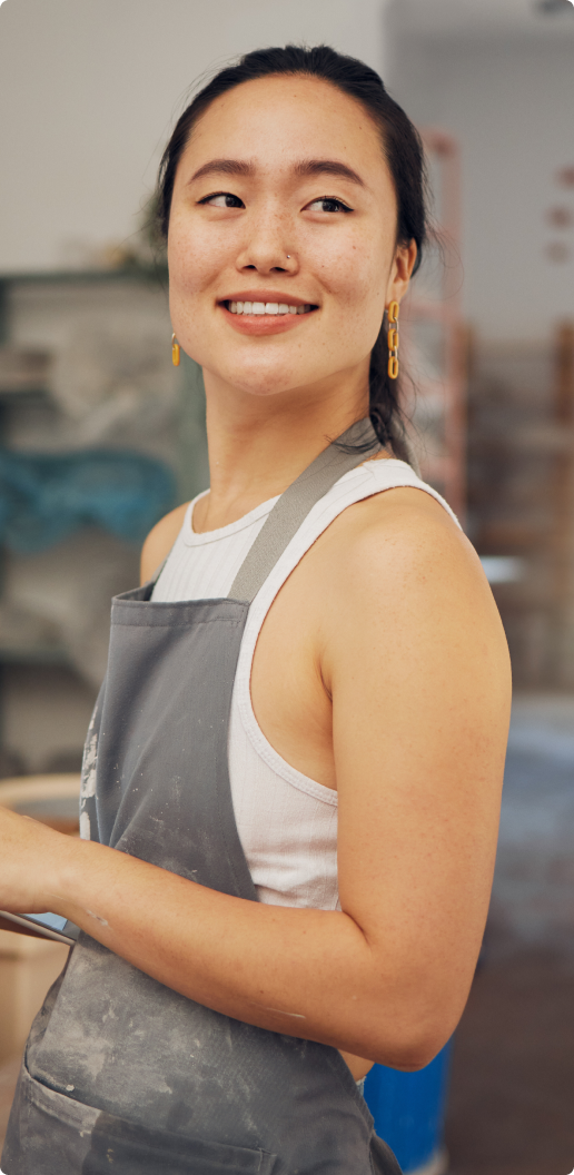 Smiling asian woman wearing a white tank top and a gray apron holding a tablet in a pottery workshop.