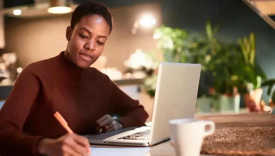 A woman sits in a cafe and takes notes next to her laptop.