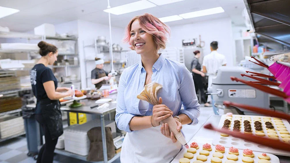 Smiling woman in a bakery kitchen holding a piping bag wearing a blue button down shirt and a white apron.