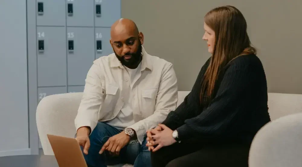  A man with a beard wearing a white button-down shirt points at a laptop screen while sitting on a curved couch and discussing something with a woman in a dark sweater in an office setting.