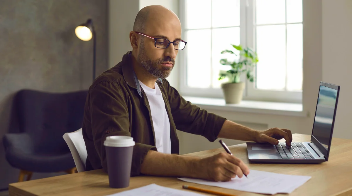 Bald man with a beard sits taking notes at his desk