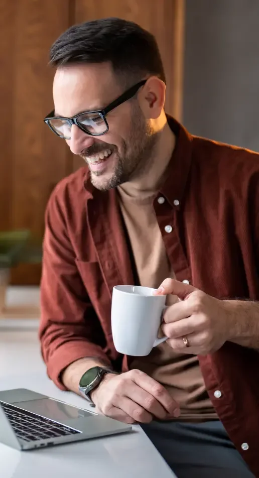 A man with glasses and a red button up shirt drinks coffee and smiles at his laptop.