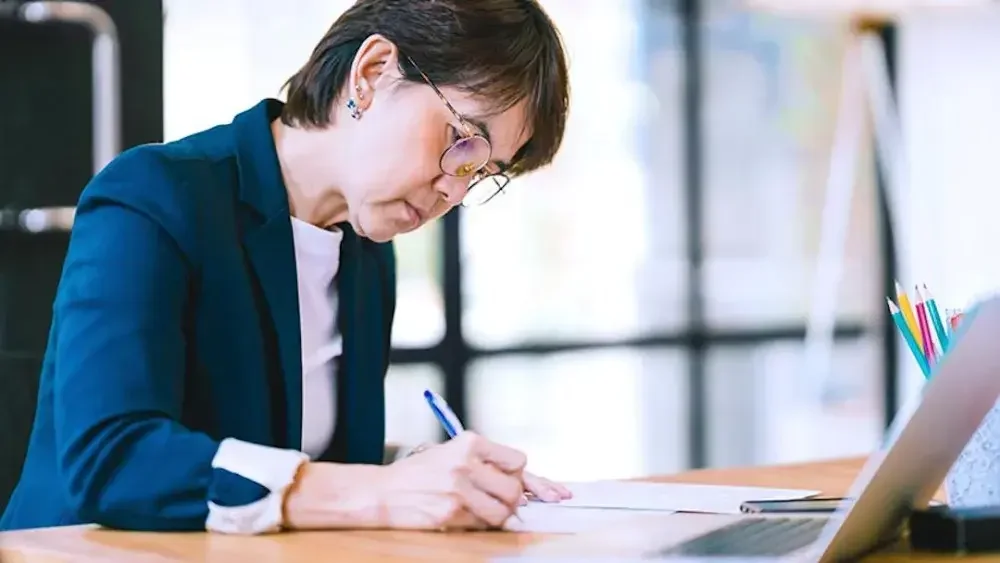 Woman writing on paper on a desk in front of laptop