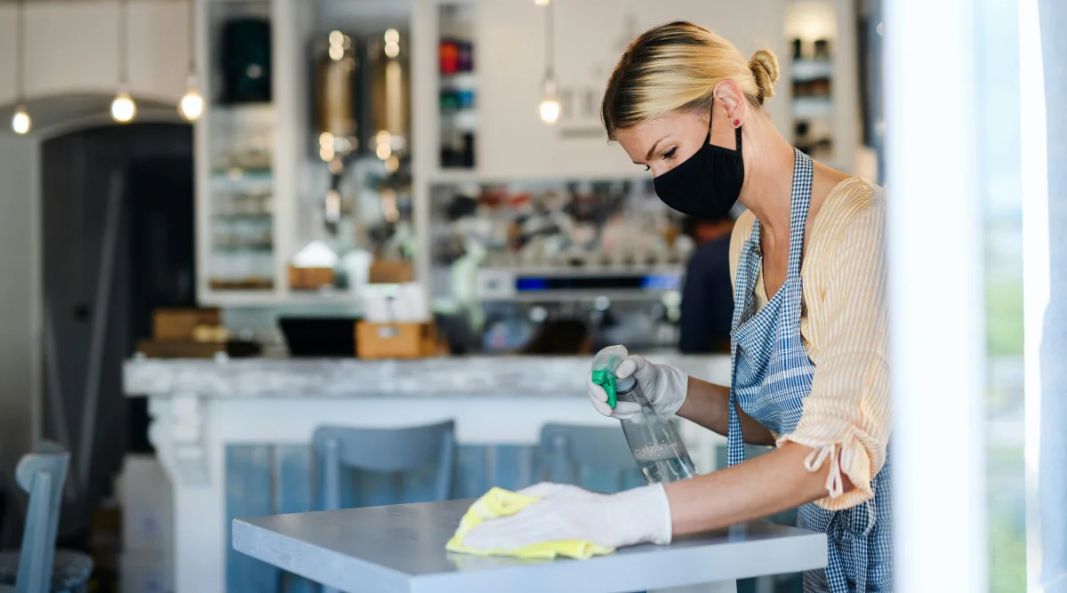 A blonde woman cleans a kitchen countertop while wearing gloves, an apron, and a protective face mask.
