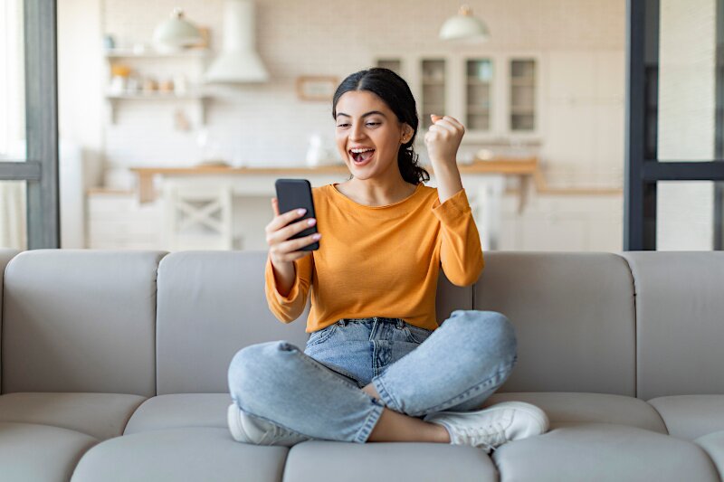 A young woman is sitting on a sofa looking at her phone very excitedly. 