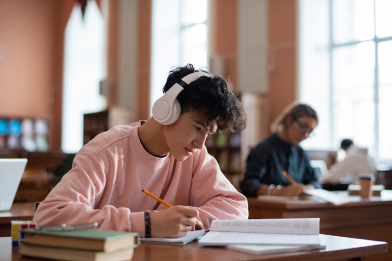 A young man with headphones sits at a desk doing coursework.