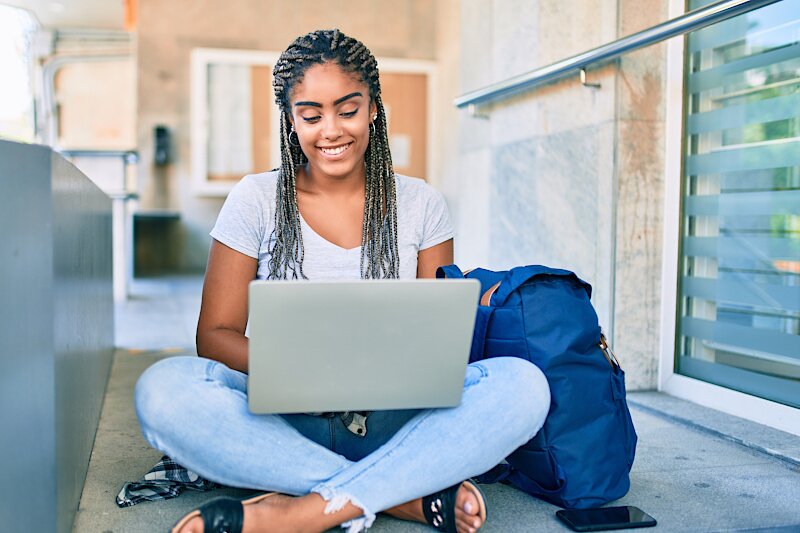 A young woman is sitting outside on her laptop smiling. 