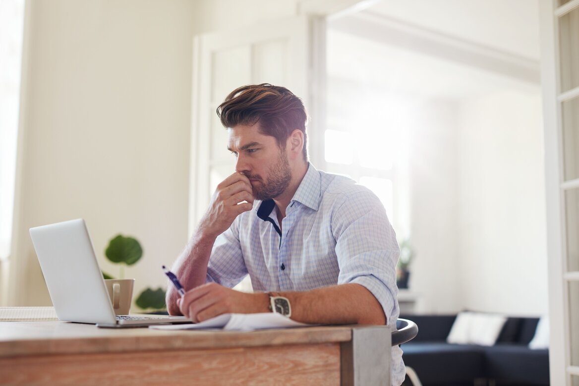 Person sitting at a desk with a laptop, thinking carefully while making notes about their tax return