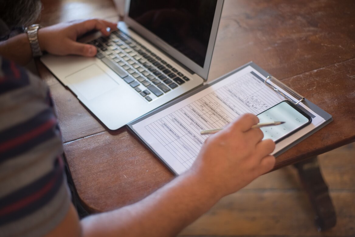 Man filling in a paper tax form while using a laptop and calculator app on his phone