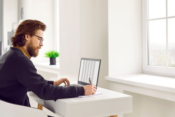 Man wearing glasses sits at a desk writing notes while looking at a laptop displaying a spreadsheet.