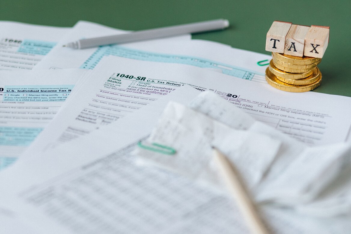 Tax return forms with wooden blocks spelling TAX stacked on coins, representing the cost of self assessment