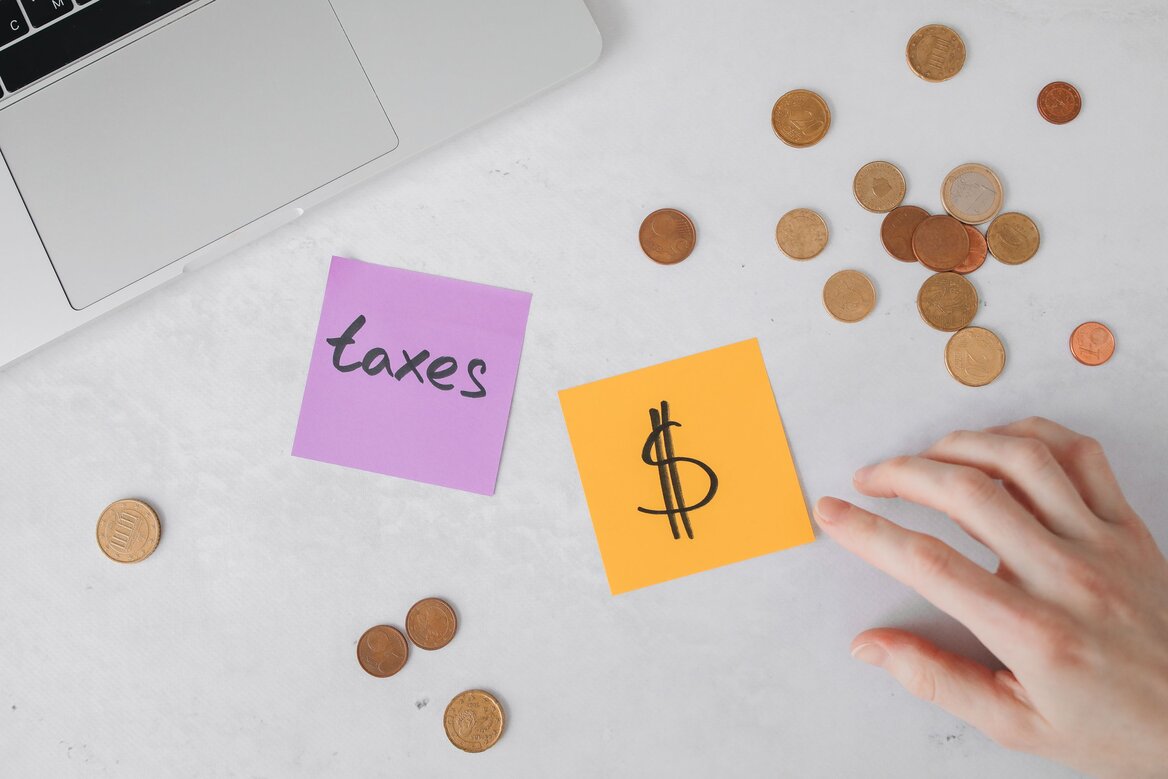 Sticky notes reading "taxes" and a dollar sign next to coins and a laptop, symbolising budgeting for a tax bill