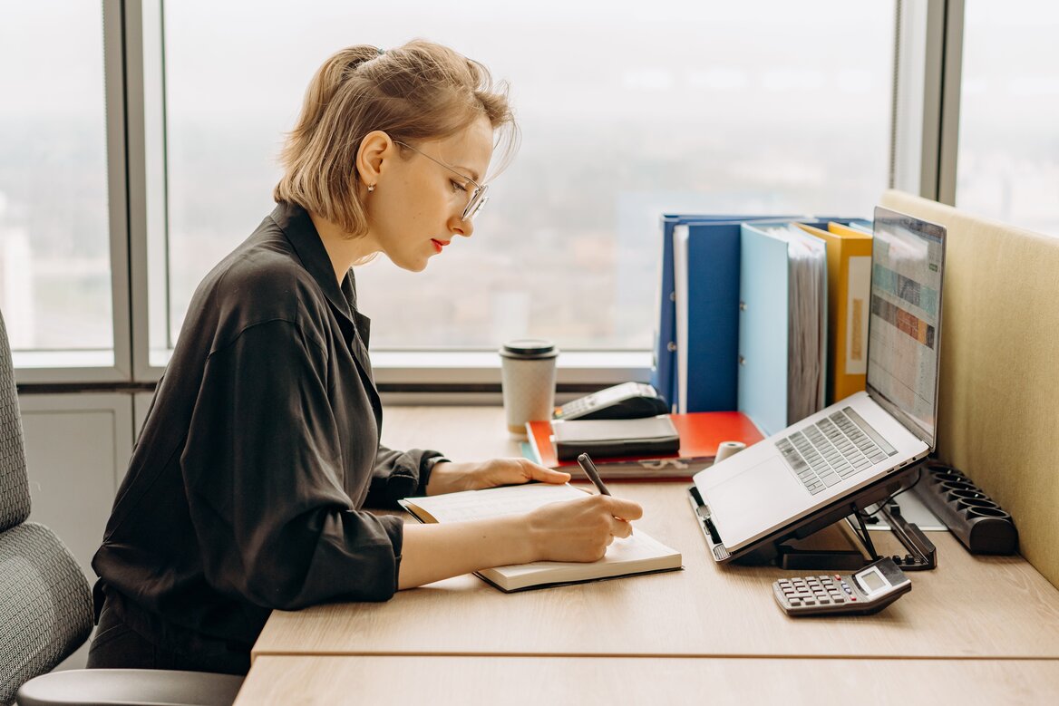 Professional woman writing in a notebook at an office desk with a laptop and documents, reviewing why HMRC’s own record matters separately from your MTD obligations.