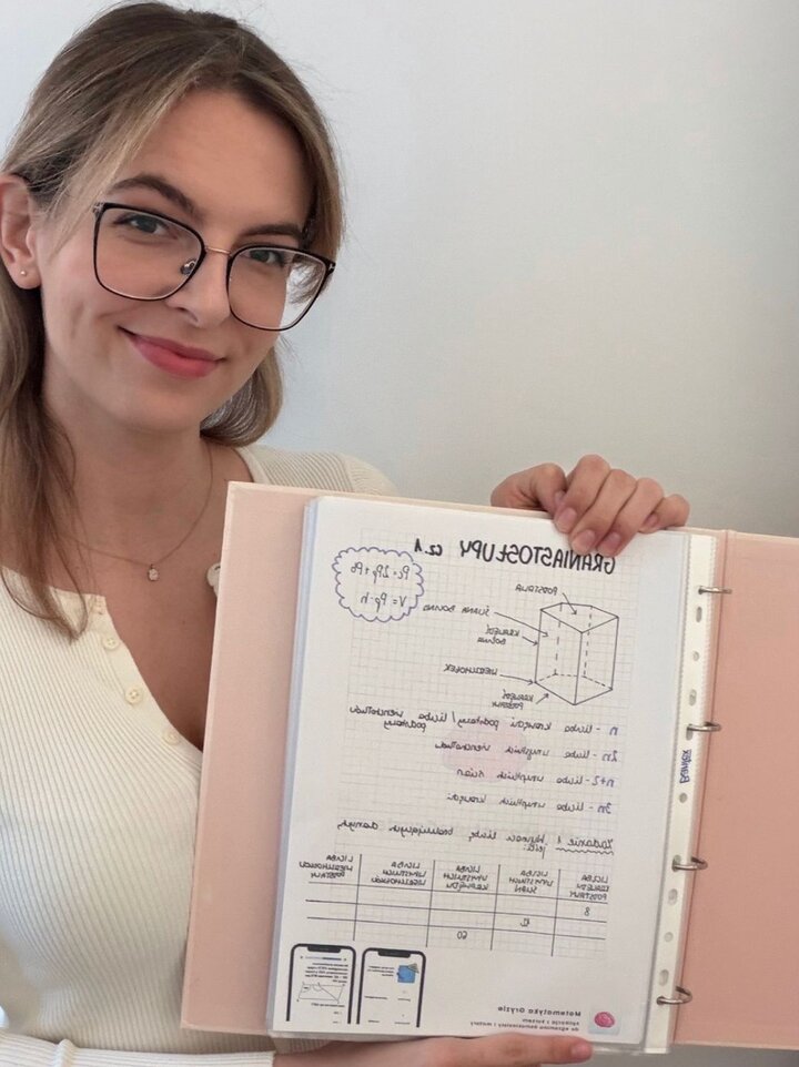 Zdjęcie przedstawia młodą kobietę o jasnej karnacji i blond włosach, noszącą okulary w ciemnych oprawkach. Kobieta uśmiecha się lekko do aparatu, trzymając przed sobą otwarty, różowy segregator z notatkami.

Kobieta ma na sobie biały, prążkowany kardigan z guzikami.

Nosi delikatny złoty naszyjnik z małą zawieszką.

Jej włosy są upięte lub zaczesane do tyłu, z kilkoma luźnymi pasmami okalającymi twarz.

W segregatorze znajduje się kartka w kratkę z odręcznymi notatkami w języku polskim, zatytułowana na górze dużymi literami: „GRANIASTOSŁUPY cz. 1”.

Na środku widnieje rysunek techniczny prostopadłościanu z zaznaczonymi i podpisanymi elementami (np. krawędź boczna, wierzchołek, podstawa).

W lewym górnym rogu, w ozdobnej chmurce, zapisano wzory matematyczne: Pc=2Pp+Pb oraz V=Pp⋅h.

Poniżej rysunku znajdują się definicje oznaczeń (np. n – liczba wierzchołków podstawy) oraz tabela z danymi dotyczącymi różnych graniastosłupów.

Na samym dole strony widać dwie małe grafiki przypominające ekrany smartfonów z dodatkowymi ilustracjami geometrycznymi.

Tło jest jednolite, jasne i neutralne, co skupia całą uwagę na osobie i prezentowanych przez nią materiałach edukacyjnych.