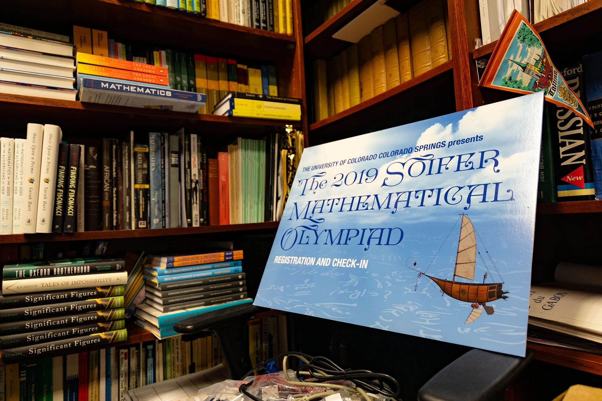 A poster from the 2019 Soifer Mathematical Olympiad sits surrounded by Soifer’s dense office library. Photo: Chase McCleary, Rocky Mountain PBS