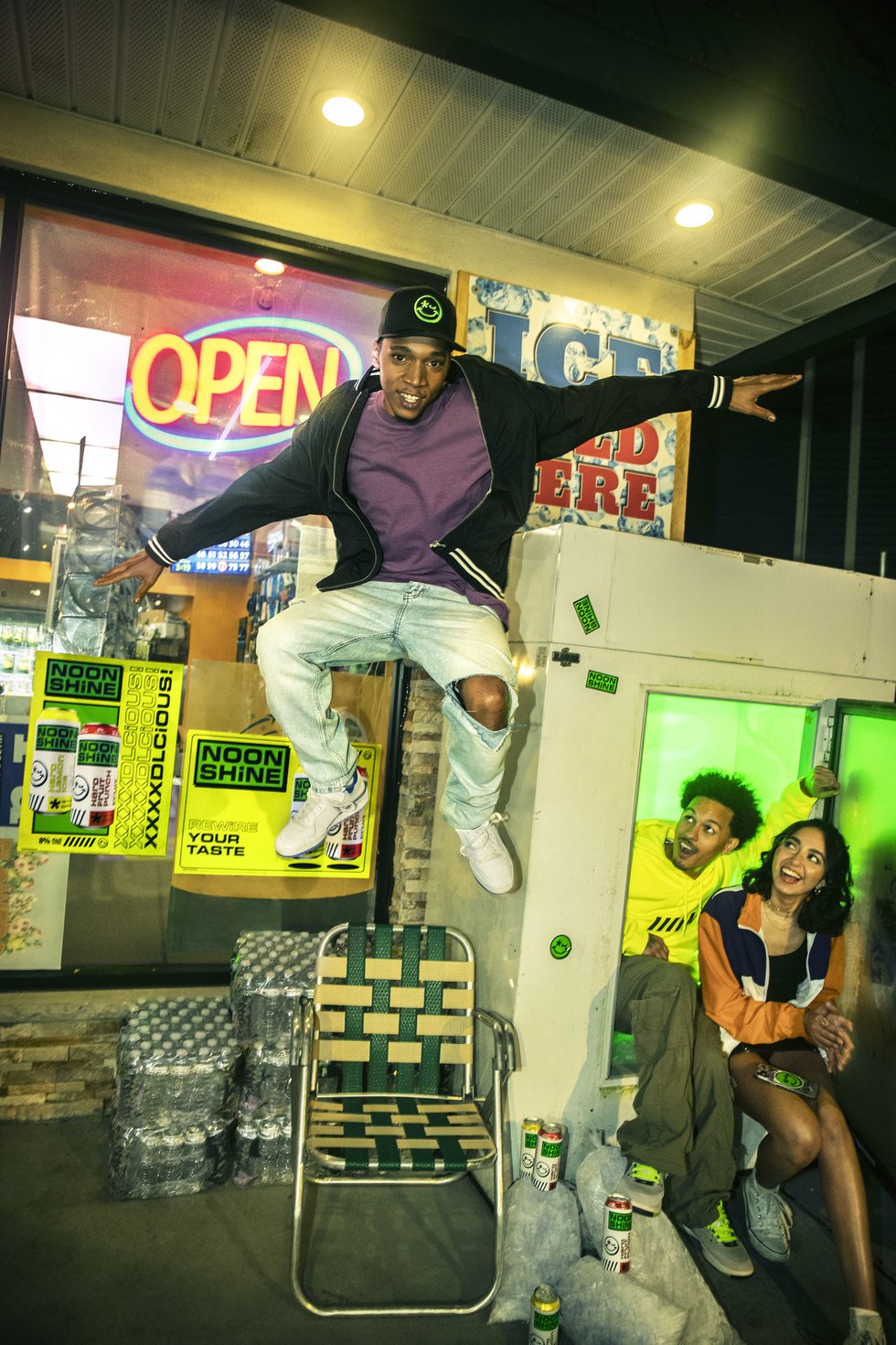 A man enthusiastically jumping in front of a convenience store adorned with Noonshine branding. The store features cans of Noonshine and a man and a woman are seen sitting and enjoying their drinks. inside a commercial ice chest.