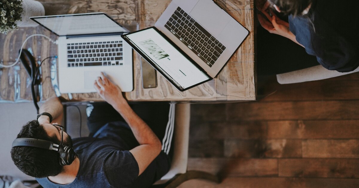 Two professionals with laptops sit at a shared desk comparing prospecting notes