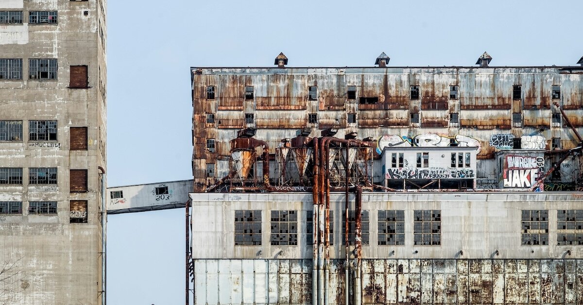 A dilapidated building with grafitti stands adjacent to another older, concrete building