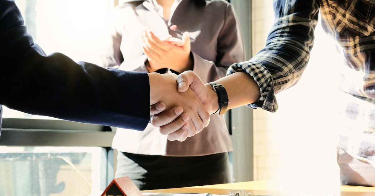 Two professionals shake hands as another person looks on with clasped hands