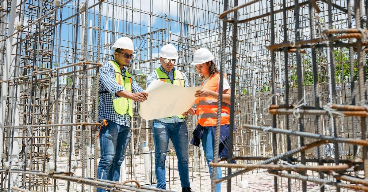 Three people review construction plans wearing hard hats at a development site.