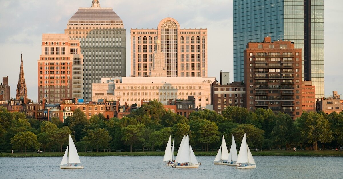 Boats in the Boston harbor float in front of a park and retail buildings