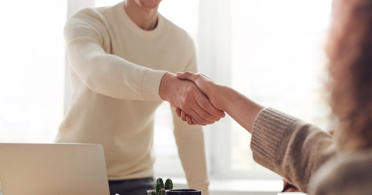 A masc in a cream sweater and femme in an oatmeal sweater shake hands over a desk.