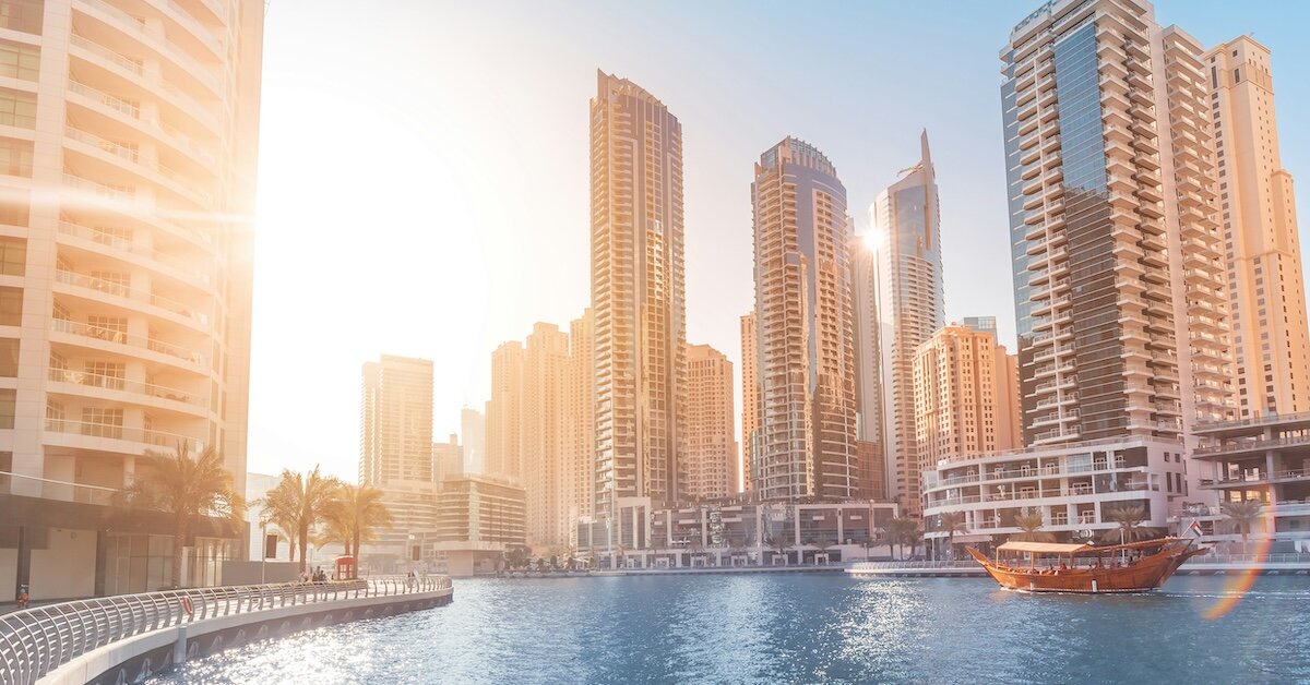 The sun shines behind several shiny commercial buildings with a bay and a floating gondola in the foreground