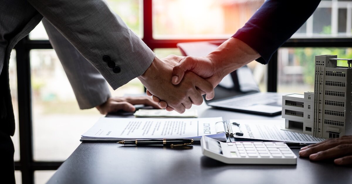 Two men shaking hands over a model commercial building, a contract and a calculator