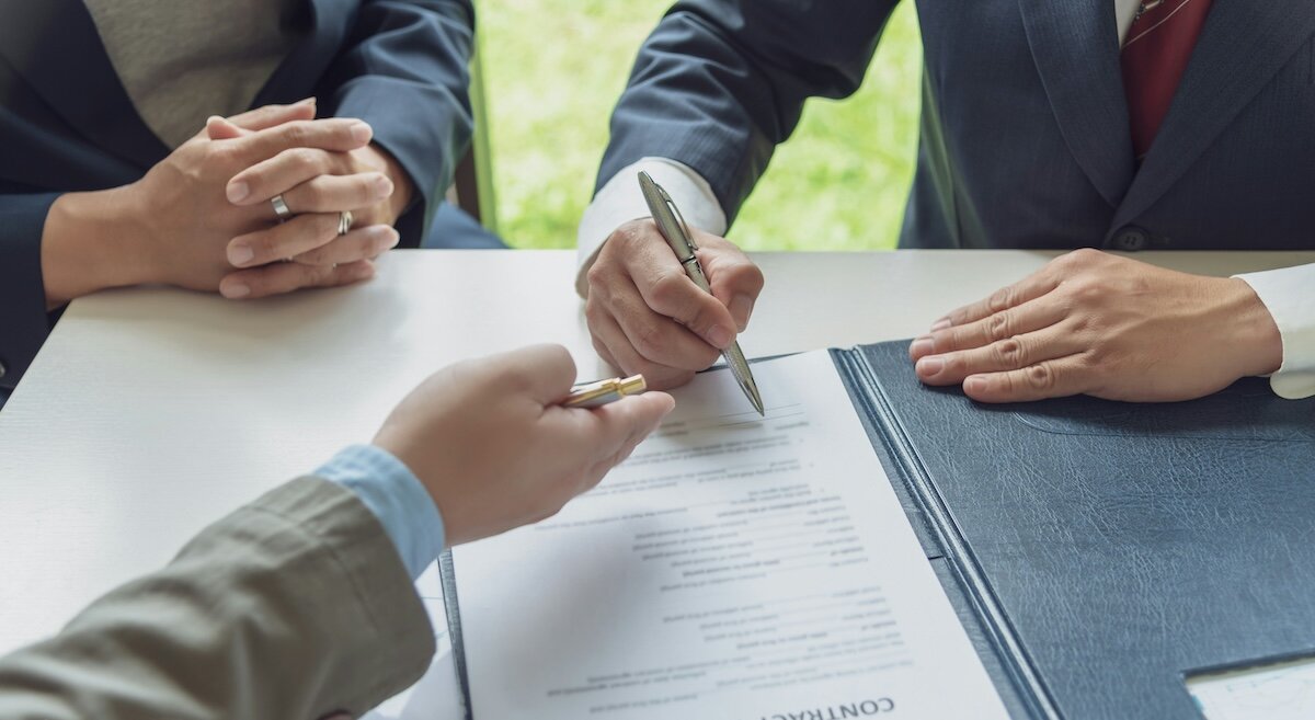 Three people sit at a table with a contract open, one poised to sign on the dotted line