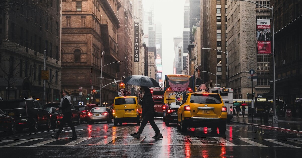 A Manhattan street in the rain with pedestrians crossing in front of taxis while holding umbrellas
