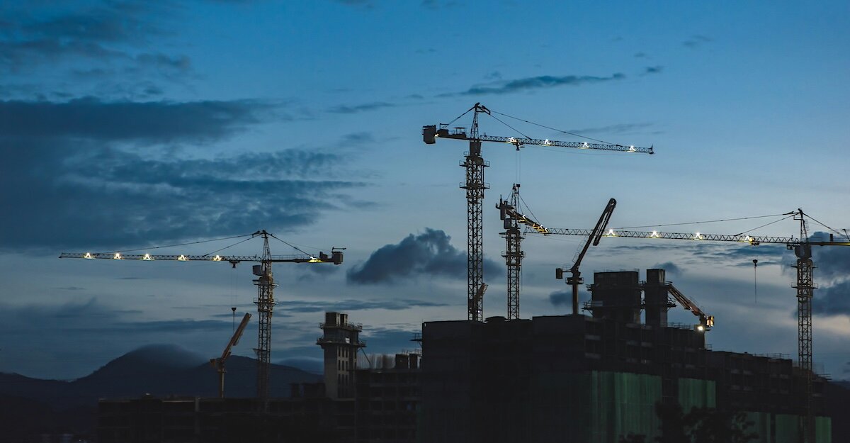 Construction cranes with lights stand in shadow against the twilight sky above commercial buildings