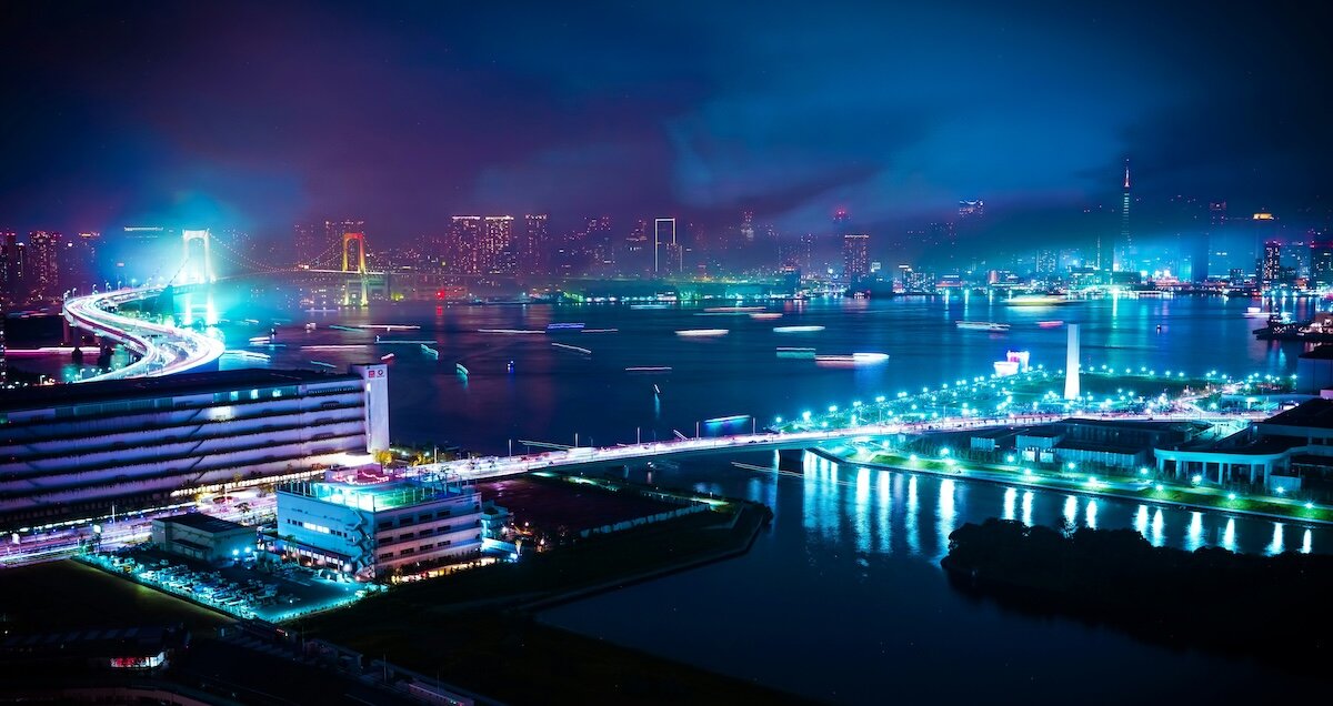 A futuristic city at night with rainbow colored lights illuminating the buildings on the bay