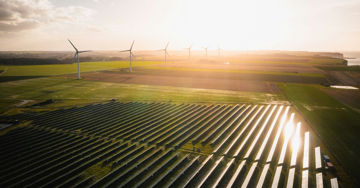 A solar farm with sunshine beaming down and wind turbines in the background